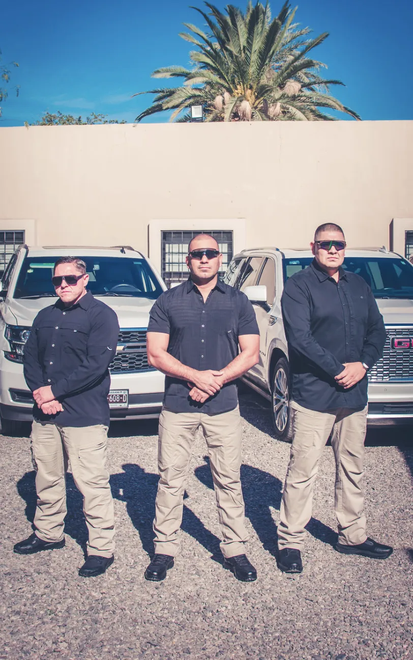 Tres guardias de seguridad privada con gafas oscuras y uniforme negro, de pie frente a vehículos blindados en un entorno seguro y profesional.