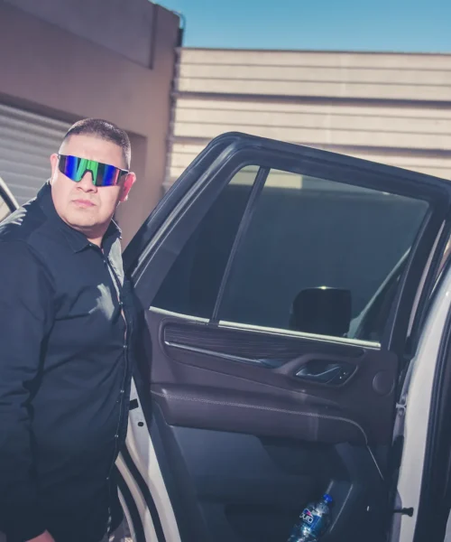 Escolta de seguridad privada con gafas de sol llamando por radio junto a una camioneta blindada.