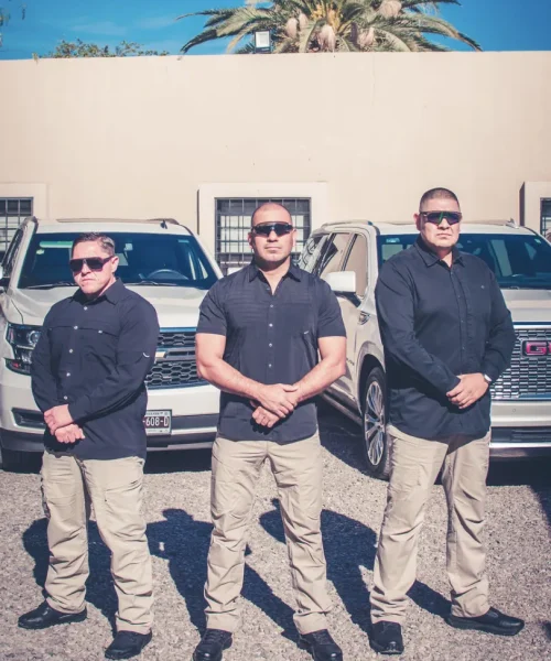 Tres guardias de seguridad privada con gafas oscuras y uniforme negro, de pie frente a vehículos blindados en un entorno seguro y profesional.